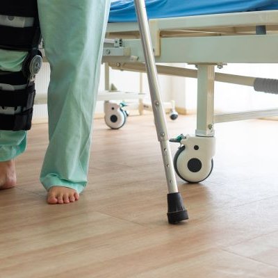 The legs of a person by a hospital bed in green clothing with a braced knee and crutches